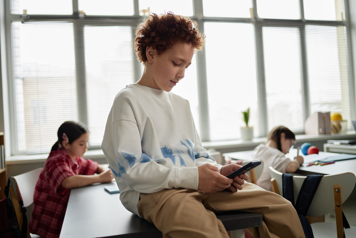 Caucasian Boy Using Smartphone While Sitting on Desk in Classroom Setting